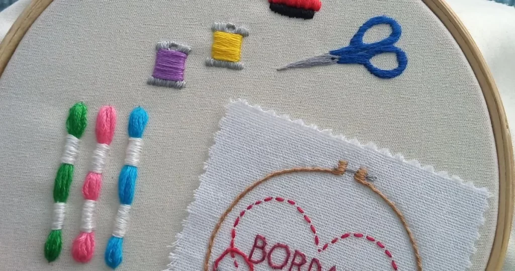 tela bordada em bastidor com desenho de linhas meadas, agulhas e alfinetes, tesoura e uma mini tela bordada com um coração escrito bordado. peça produzida pela artesã andrea catão de rosália rosarinha e agulhas da resistência