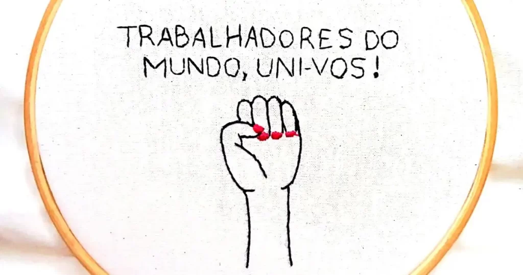 uma mão feminina em punho e a frase "trabalhadores do mundo, uni-vos!" bordada em um tecido de algodão e emoldurada em um bastidor de madeira. peça da artesã andrea catão de rosália rosarinha e agulhas da resistência