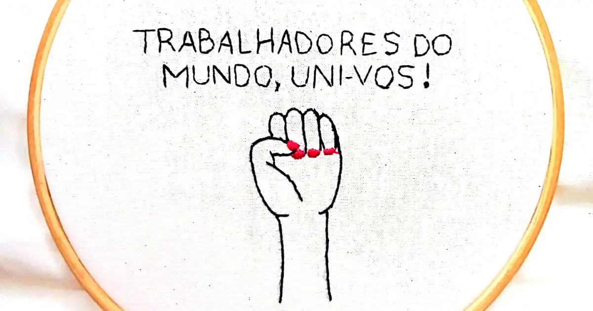 uma mão feminina em punho e a frase "trabalhadores do mundo, uni-vos!" bordada em um tecido de algodão e emoldurada em um bastidor de madeira. peça da artesã andrea catão de rosália rosarinha e agulhas da resistência