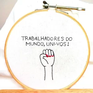 uma mão feminina em punho e a frase "trabalhadores do mundo, uni-vos!" bordada em um tecido de algodão e emoldurada em um bastidor de madeira. peça da artesã andrea catão de rosália rosarinha e agulhas da resistência