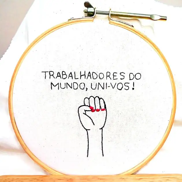 trabalhadores interna uma mão feminina em punho e a frase "trabalhadores do mundo, uni-vos!" bordada em um tecido de algodão e emoldurada em um bastidor de madeira. peça da artesã andrea catão de rosália rosarinha e agulhas da resistência