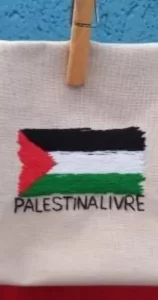 bandeira da palestina bordada sobre um tecido de algodão preso a um pregador de roupas. no bordado, também está escrito "palestina livre" abaixo da bandeira. a peça é da artesã andrea catão