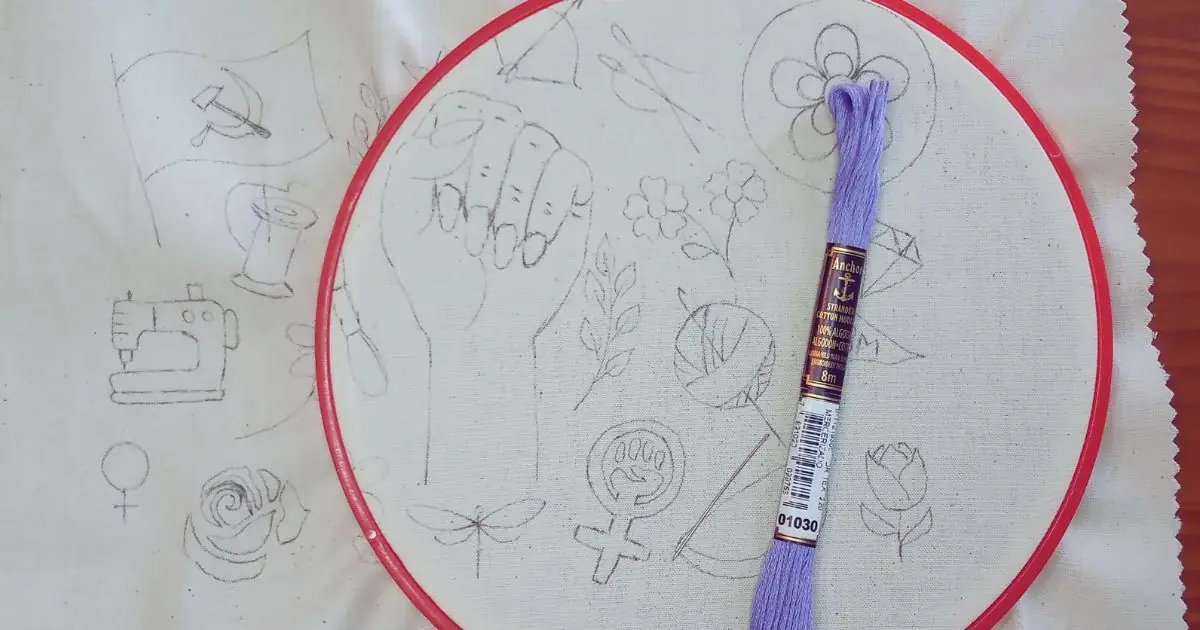 tecido de algodão já riscado com um desenho a ser bordado e seguro por um bastidor redondo de plástico, que tenciona o tecido para bordar e uma linha meada na cor roxa sobre o tecido