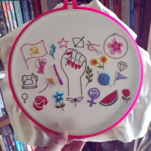 bordado em tecido de algodão em um bastidor de plástico com vários símbolos para o banner do site agulhas da resistência para a série manifesto bordado, criado e produzido pela artesã andrea catão. são bordados de desenhos de um punho feminino erguido, flores, folhas, como rosas e margarida, espelho de vênus, símbolo feminista, bandeira comunista, novelo de linha, carretel, agulhas, libélula, estrela etc