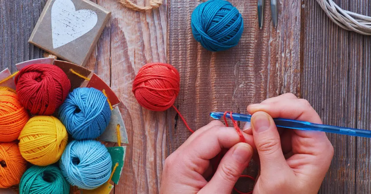 mãos segurando uma agulha de crochê fazendo crochê com um novelo de linha vermelha. mãos sobre uma mesa de madeira e ao lado uma tigela com novelos de linha coloridos. a imagem ilustra o texto "Pinterest, não bote IA no meu artesanato" sobre uso de imagens de inteligência artificial como se tivessem sido produzidas por mãos humanas. texto de andrea catão para o site agulhas da resistência